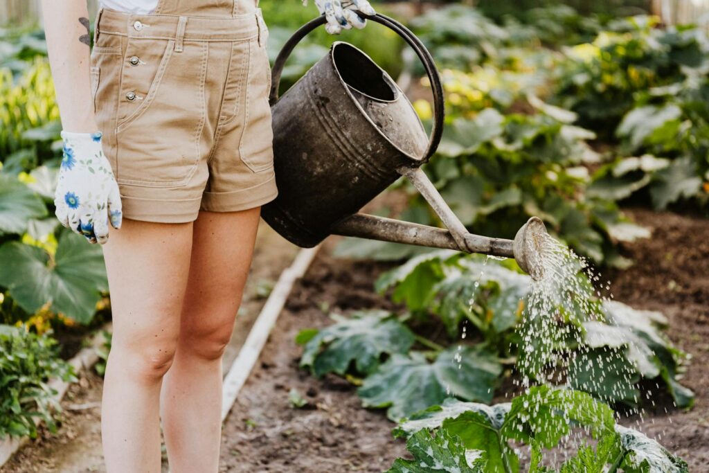 A woman waters plants in a lush home garden with a metal watering can.